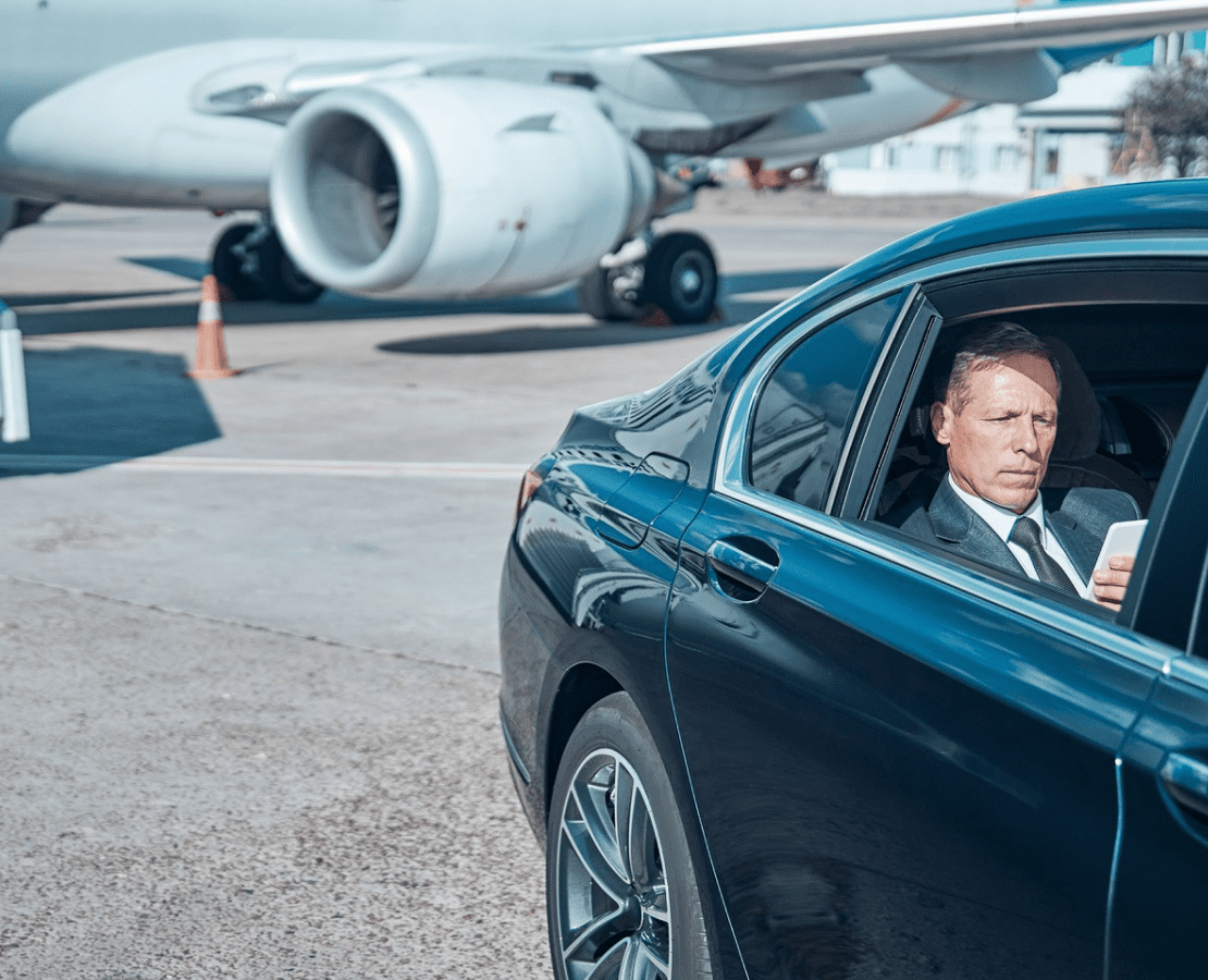 Well-dressed man in dark SUV at airport tarmac with commercial aircraft in background.