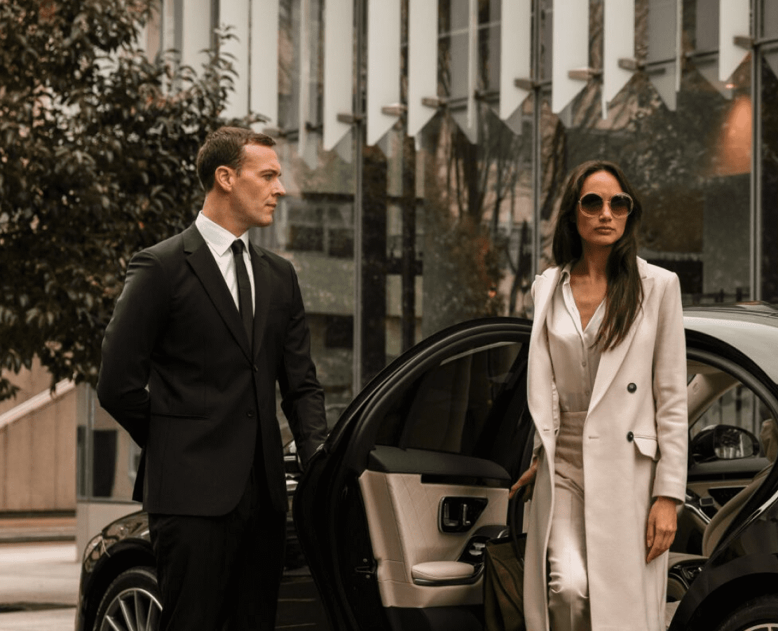 Two elegantly dressed professionals stand beside a luxury SUV in an upscale urban setting.